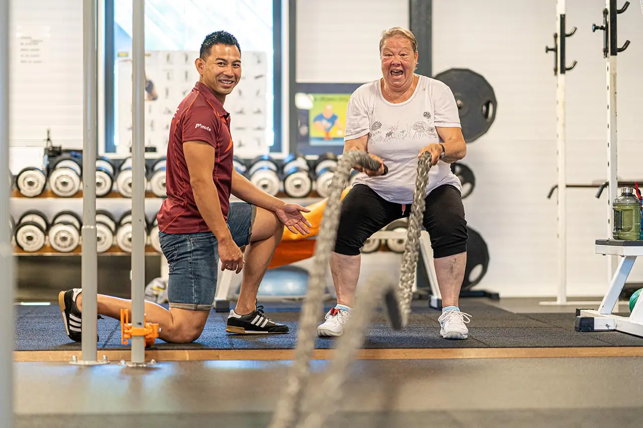 Sporter lacht tijdens een oefening met battle ropes, instructeur moedigt aan